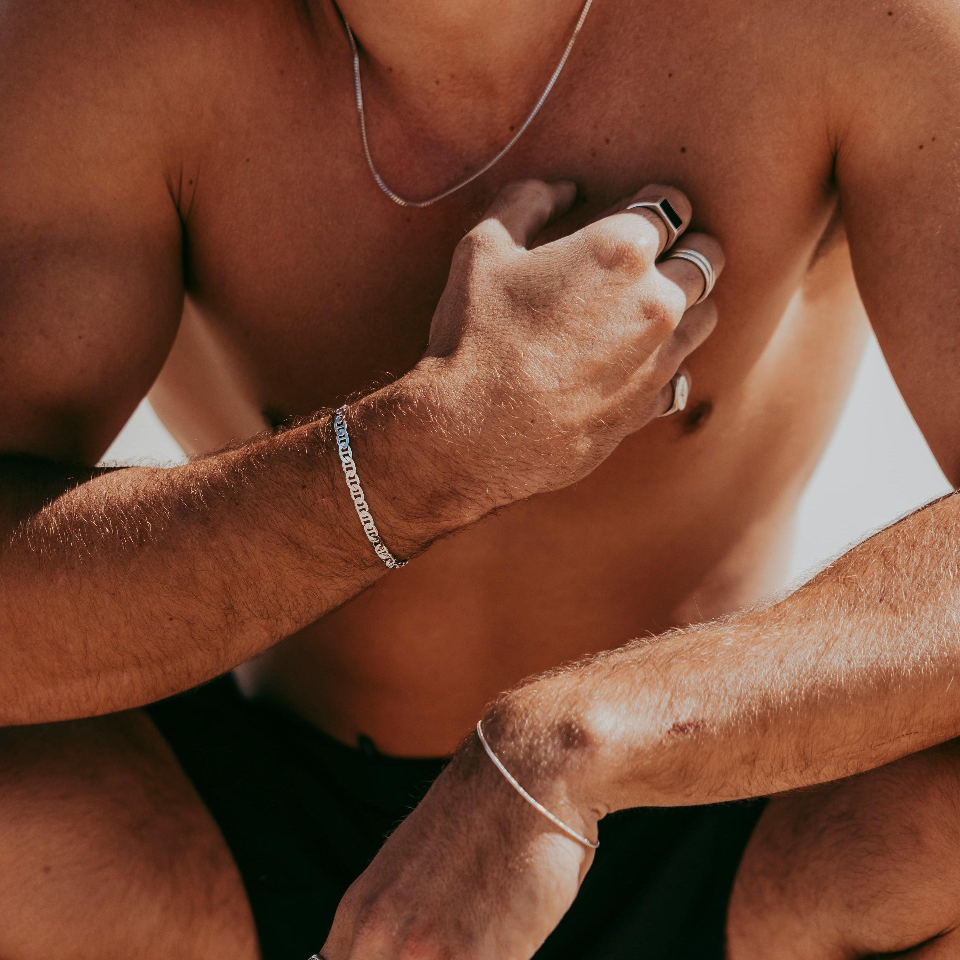 A shirtless man wearing multiple silver rings, bracelets, and a necklace, crouches with his hand on his chest and the other resting on his knee. The background is bright and sunlit.