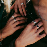 Close-up of two people wearing silver rings and necklaces, gently touching each others hands and chest, dressed in dark shirts with sunlight highlighting their skin and jewelry. v2