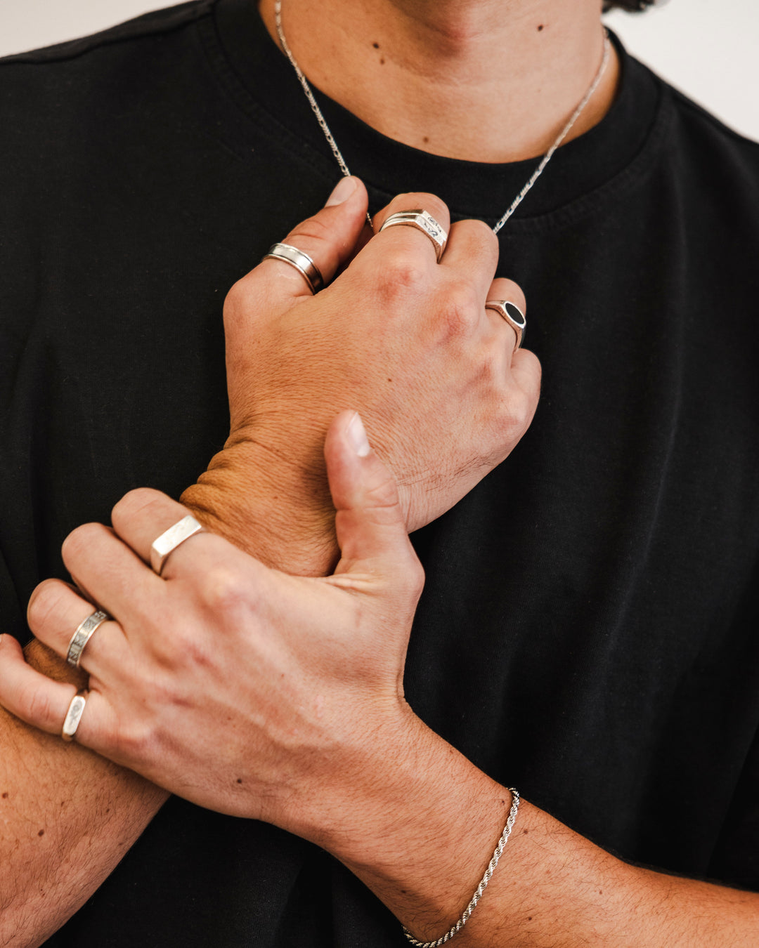 A person in a black t-shirt holds their wrist, showing multiple silver rings on their fingers, a chain bracelet, and a necklace. Only the hands, part of the neck, and torso are visible.
