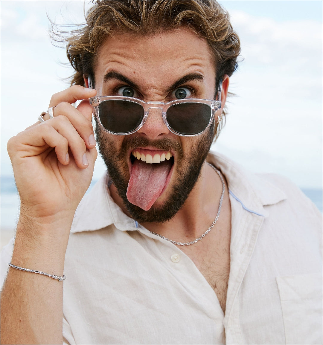 A bearded man wearing sunglasses and a white shirt sticks out his tongue playfully while looking at the camera on a beach.