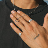 Person wearing a necklace and two rings with a tattoo on their hand, outdoors.