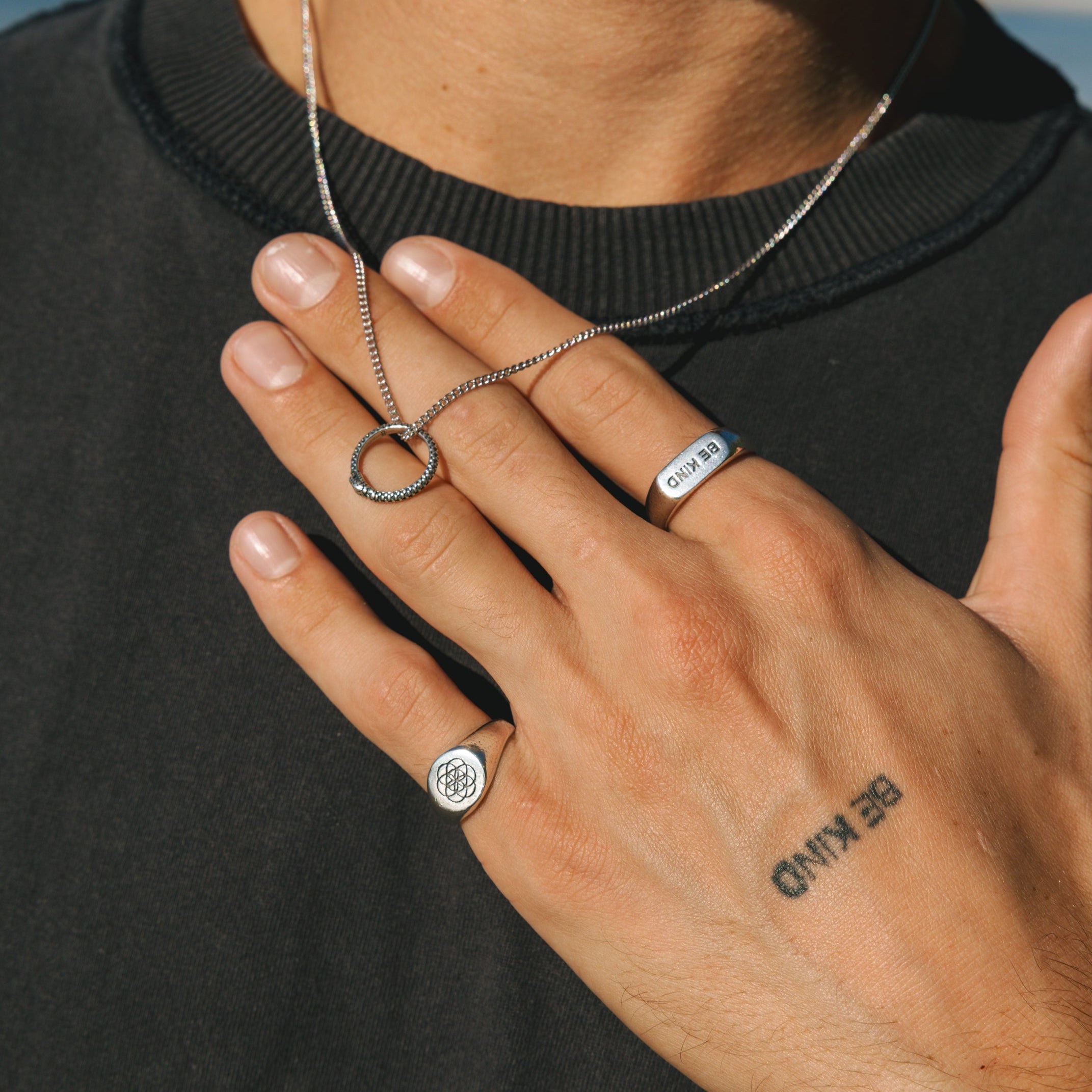 Person wearing a necklace and two rings with a tattoo on their hand, outdoors.