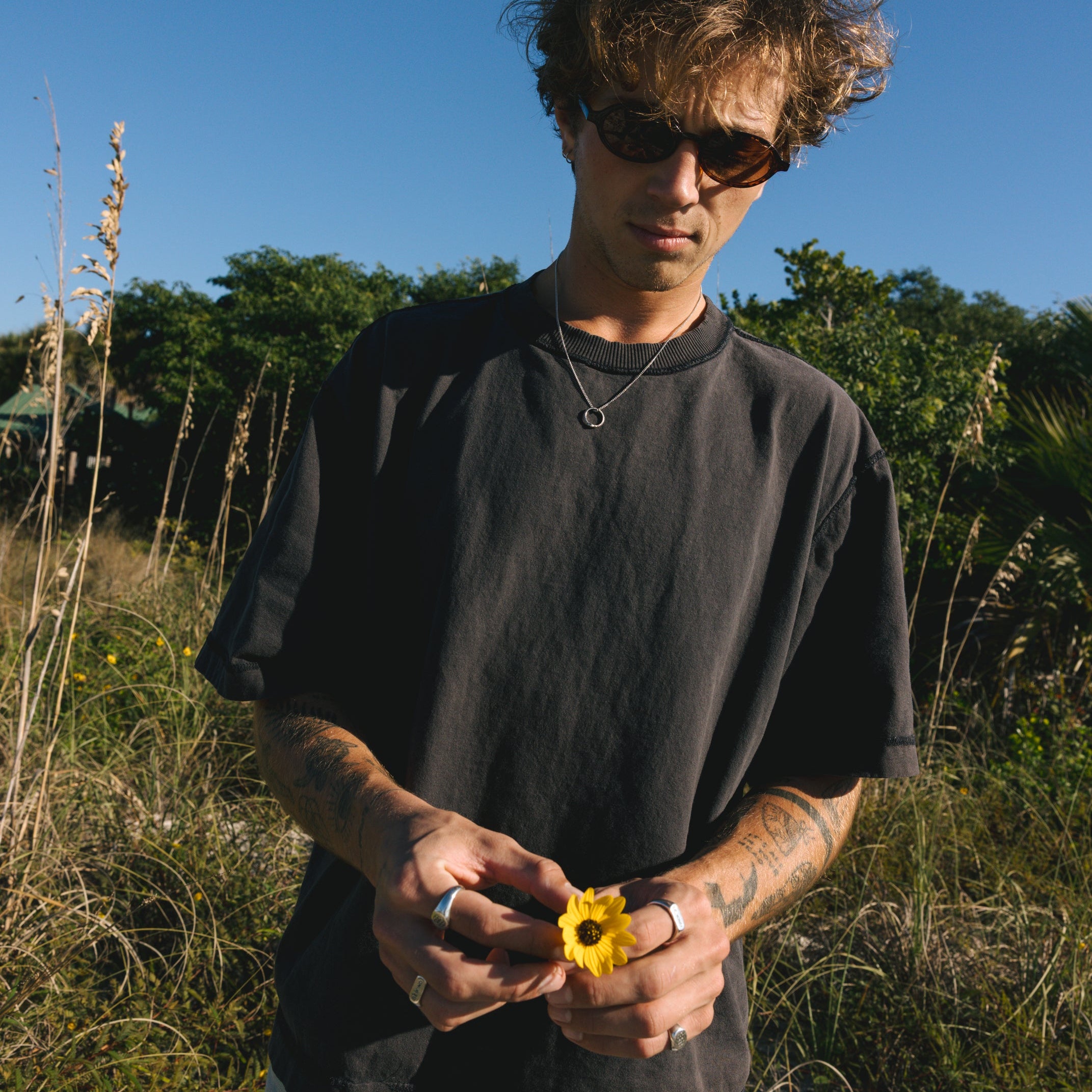 Man holding a yellow flower in a natural setting with trees and blue sky wearing men's jewellery.