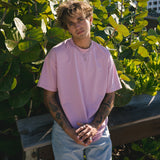 Person wearing a pink shirt and light blue jeans standing in front of green foliage with a building in the background wearing men's jewellery.